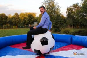 A person in a blue shirt sits on a large inflatable football ball, surrounded by a colorful inflatable barrier in a grassy outdoor setting.