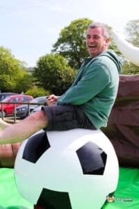 A person sits on a large inflatable football ball in a sunny outdoor setting, surrounded by greenery and parked cars in the background.