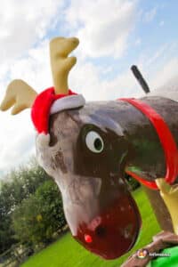 A festive inflatable reindeer with antlers and a red Santa hat, set against a cloudy sky and green grass backdrop.
