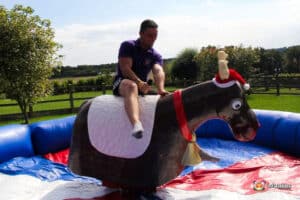 A person rides a colorful inflatable bull in a sunny outdoor setting, surrounded by greenery and a blue inflatable safety border.