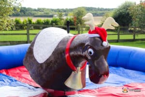 A festive inflatable bull ride shaped like a reindeer, adorned with a Santa hat and bells, set in a colorful arena outdoors.