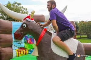 A person in a purple shirt rides an inflatable reindeer decorated with a Santa hat, beside a colourful winter-themed backdrop.