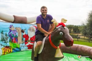 A person rides a festive inflatable reindeer, surrounded by a colorful backdrop featuring Santa and elves in a holiday scene.