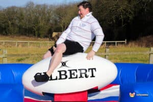 A person balances on a large inflatable rugby ball, set against a sunny outdoor background with green trees.