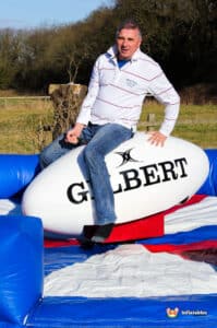 A person sits on a large inflatable rugby ball, surrounded by a colorful padded surface, enjoying a fun outdoor activity.