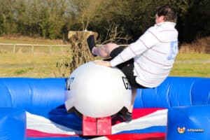A person attempts to stay on a mechanical bull, surrounded by a colorful inflatable ring, in a green outdoor setting.