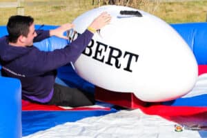 A person is positioned on an inflatable bucking rugby ball, leaning back as they prepare to stay balanced on the ride. Brightly colored mat beneath.