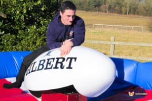 A person is riding a large inflatable rugby ball on a colourful inflatable surface, with a scenic park background.