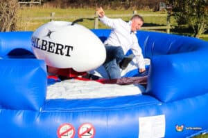 A person on an inflatable bull riding attraction, wearing a white shirt, attempts to maintain balance while leaning sideways.