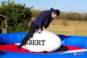 A person in a purple hoodie balances on a large inflatable rugby ball in a colorful inflatable play area, with a grassy field in the background.
