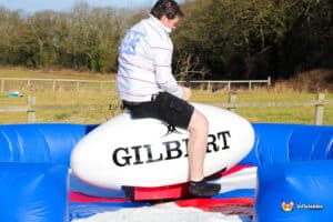 A person balances on a large inflatable rugby ball surrounded by a colorful safety mat in an outdoor setting.