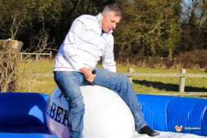 A person rides a mechanical bull, balancing on a large inflatable surface surrounded by green grass and trees in the background.