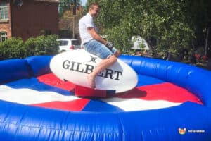 A person sits on a large inflatable rugby ball in a colorful bouncy castle, enjoying a sunny outdoor event.