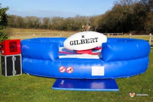 A blue inflatable rugby tackling game with a large Gilbert rugby ball and electronic score timer, set in a grassy outdoor area.