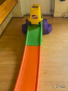 A toy car is placed on a slide in a playful children's area, surrounded by colorful structures and soft surfaces.