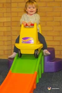 A young girl sits in a toy car, smiling as she plays in a vibrant play area designed for children.