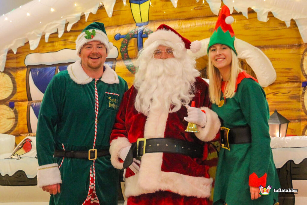 Close-up portrait of Santa Claus standing between a male and female Elf, all smiling in front of the inflatable log cabin grotto.