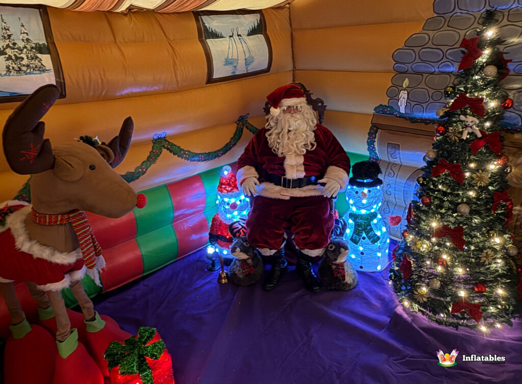 Santa Claus posing inside the inflatable grotto with a large reindeer prop, two light-up snowmen, and a lit Christmas tree in the corner.