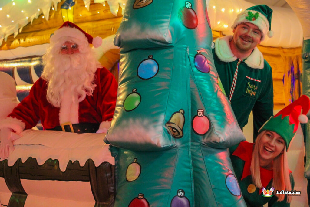 Santa Claus and two smiling Elves peeking over the top of a snow-covered inflatable barrier and a large inflatable Christmas tree.