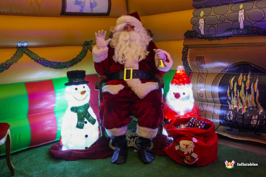 Full view of Santa Claus seated on his throne inside the grotto, waving to the camera, with a sack of gifts and light-up snowman figures flanking him by a painted fireplace.