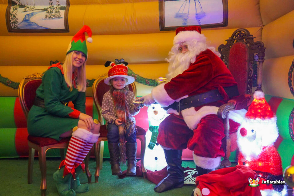 Santa Claus and an Elf seated on chairs inside the grotto, talking to a young girl in a festive hat and scarf who is seated between them.