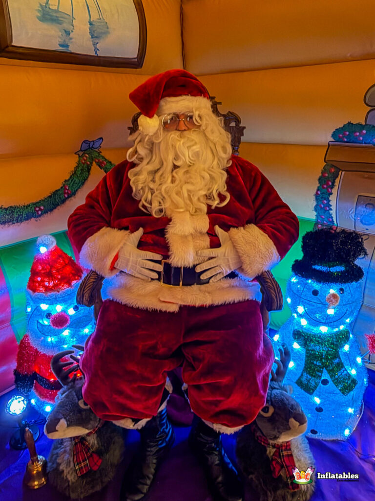 Santa Claus seated regally on his throne inside the grotto, flanked by two glowing light-up snowmen and two small plush reindeer props.