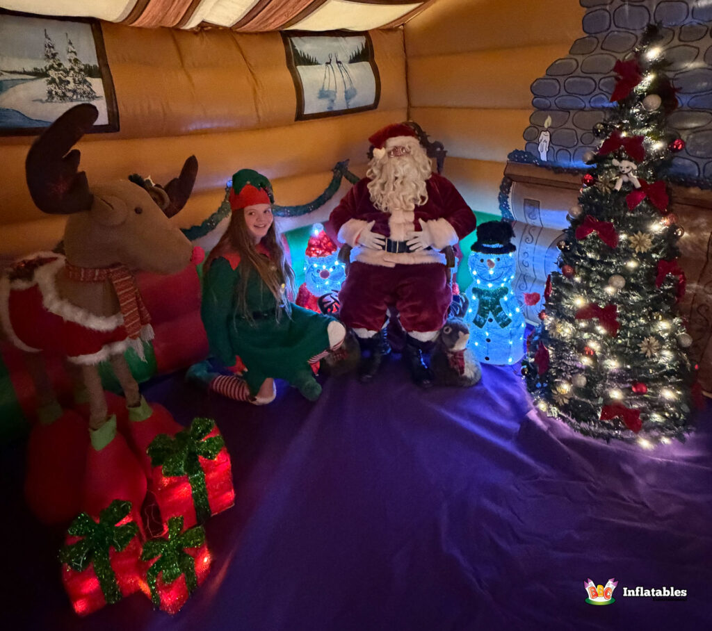 Santa and a female Elf posing inside the Christmas Grotto, sitting next to a reindeer prop, light-up decorations, and a glowing Christmas tree.