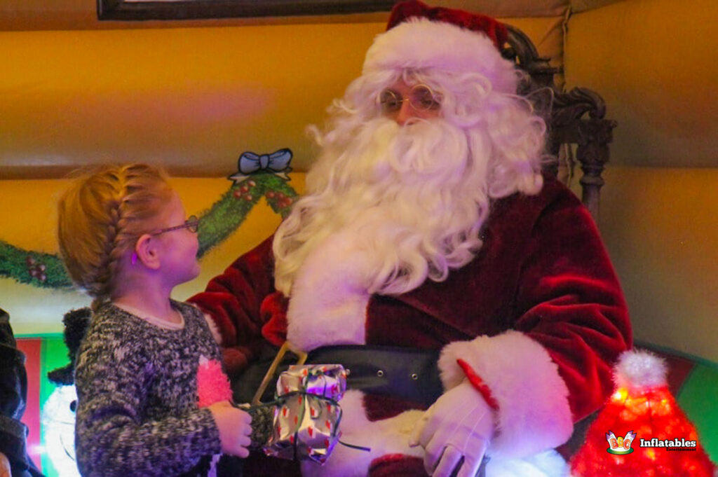 Close-up of Santa Claus seated, listening intently to a young girl who is holding a small gift, all illuminated by the warm lights of the grotto.