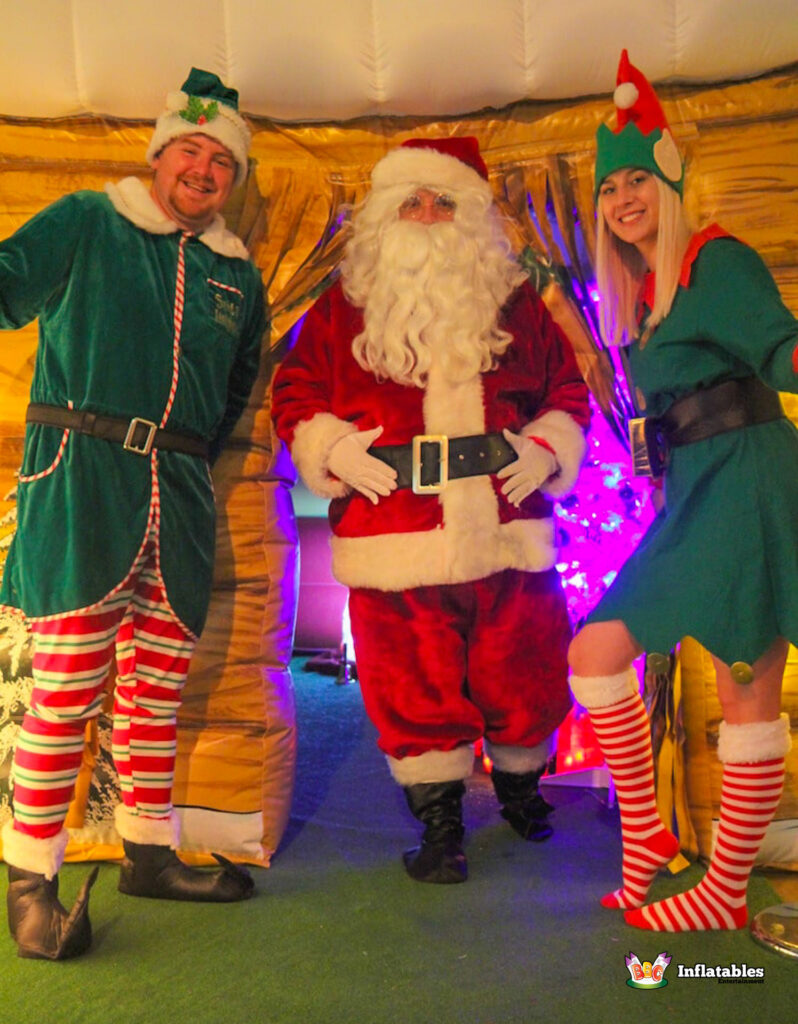Full-body shot of Santa Claus, a male Elf in striped trousers, and a female Elf in a green dress, posing together in the entrance of the inflatable grotto.