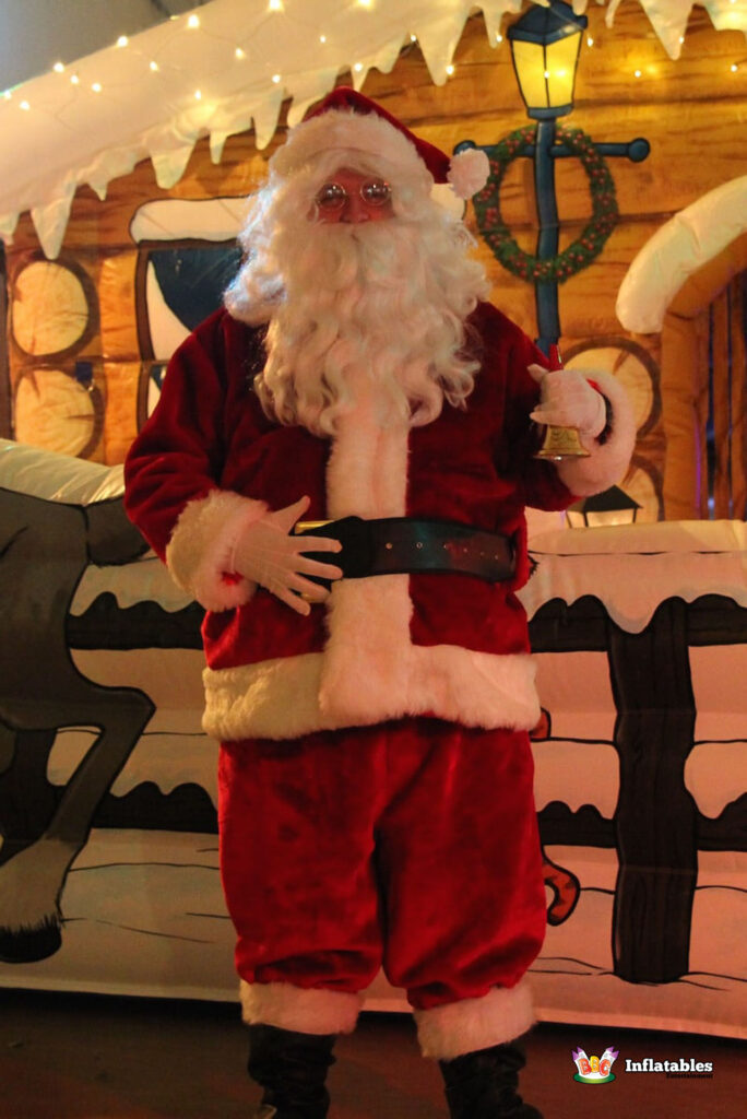 Full-length photo of Santa Claus in his velvet suit, standing in front of the log cabin grotto, holding a small gold bell and smiling.