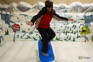 A person balances on a blue surfboard against a snowy mountain backdrop, with signs warning of avalanche dangers.