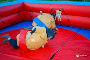 Two children wearing sumo suits play in an inflatable boxing ring on a large inflatable arena, smiling and having fun.