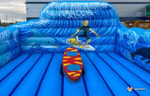A colourful inflatable surf-themed simulator with a surfer, dolphins, and wave patterns, featuring a red surfboard in the centre.