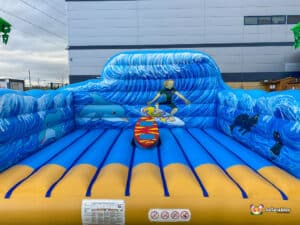 A vibrant inflatable bounce house featuring ocean waves, dolphins, a surfer, and colourful surfboard, set against a grey building backdrop.