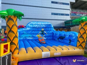 A vibrant inflatable bounce house with ocean waves, dolphins, and palm trees, featuring a surfer riding a wave, set outdoors.