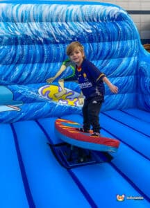 A child balances on a colorful surfboard against a backdrop of waves and a surfing graphic in a vibrant inflatable attraction.