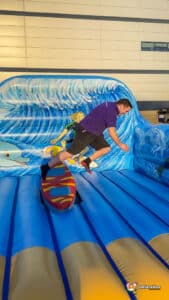 A person in a purple shirt attempts to balance on a surf simulator against a colourful inflatable backdrop of crashing waves and surfers.
