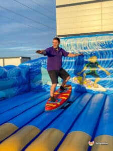 A person stands on a surfboard in a vibrant inflatable surf simulator, surrounded by colorful ocean-themed graphics.