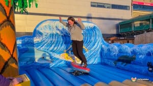 A person balances on a surfboard on an inflatable wave, surrounded by vibrant ocean-themed artwork featuring dolphins and turtles.