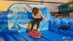 A woman balances on a surfboard in front of a colorful inflatable wave with dolphins, enjoying a fun day at a carnival attraction.