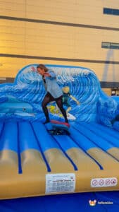 A person in a blazer balances on a skateboard atop a vibrant inflatable surf simulator platform decorated with dolphins and ocean waves.