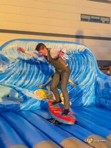 A person in a suit is surfing on an inflatable surf simulator with a colourful wave backdrop and playful dolphin design.