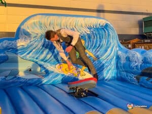 A person in a formal outfit balances on a surfboard on a colorful inflatable wave, surrounded by vibrant ocean-themed artwork.