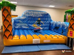 A colourful inflatable surf simulator featuring palm trees, waves, dolphins, and a surfer. Ideal for fun events.