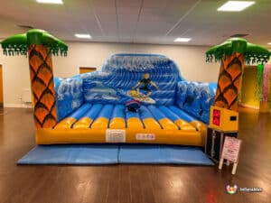 A vibrant inflatable surfing attraction with palm trees, waves, and dolphins, featuring a timer and safety instructions.