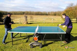 Two players are engaged in a game of table tennis outdoors, with a grassy field and trees in the background under a partly cloudy sky.