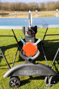 Close-up of a Kettler table tennis table with a black base and orange safety lock, set up on a grassy outdoor area.