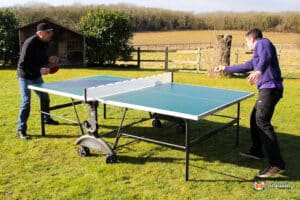 Two players engaged in a lively game of table tennis outdoors on a sunny day, with a grassy field and wooden fence in the background.