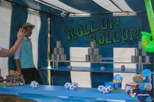 A carnival game tin can alley side stall with a vibrant blue backdrop displays stacked cans, soccer ball prizes, and colourful toys, inviting players to participate.
