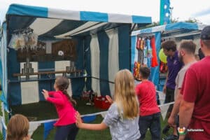 A carnival game booth with colorful tents where children aim to knock over cans with balls, while bystanders watch excitedly.